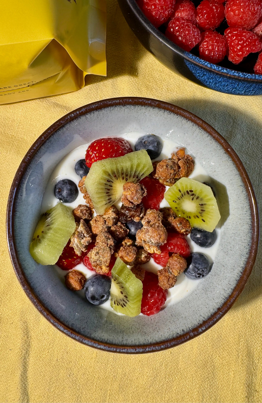 A top-down view of a bowl of Moonbeans granola with yogurt, fresh berries, blueberries, and kiwi slices, on a yellow tablecloth. A yellow bag is partially visible in the upper left, and a blue bowl of raspberries is in the upper right.