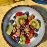 A top-down view of a bowl of Moonbeans granola with yogurt, fresh berries, blueberries, and kiwi slices, on a yellow tablecloth. A yellow bag is partially visible in the upper left, and a blue bowl of raspberries is in the upper right.