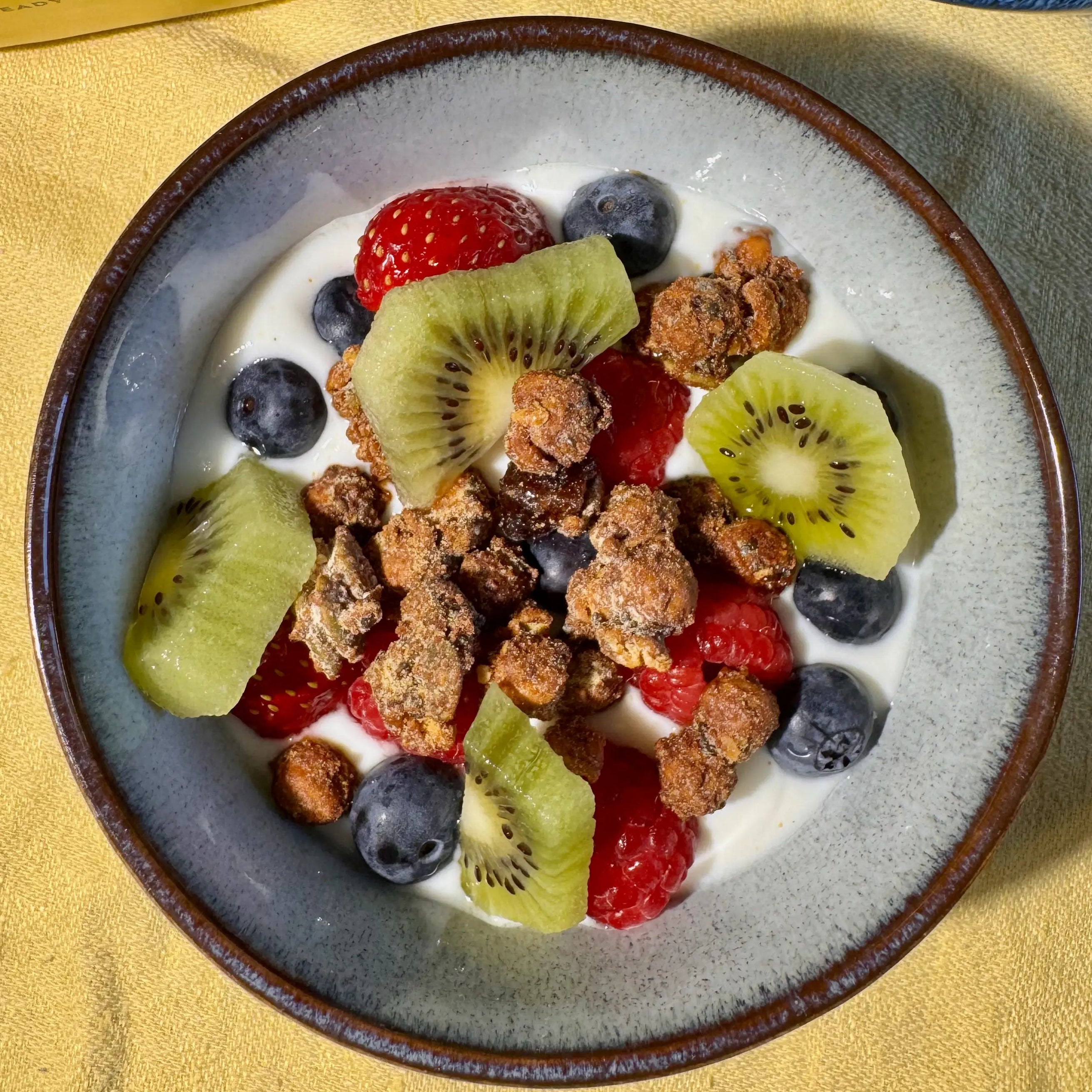 A top-down view of a bowl of Moonbeans granola with yogurt, fresh berries, blueberries, and kiwi slices, on a yellow tablecloth. A yellow bag is partially visible in the upper left, and a blue bowl of raspberries is in the upper right.