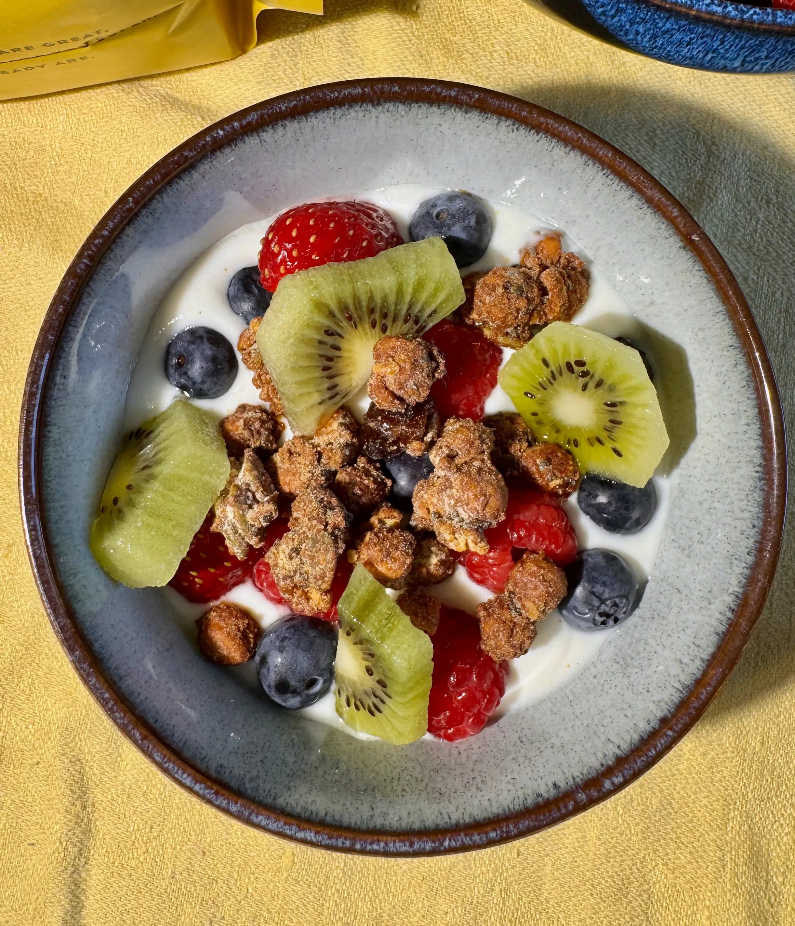 A top-down view of a bowl of Moonbeans granola with yogurt, fresh berries, blueberries, and kiwi slices, on a yellow tablecloth. A yellow bag is partially visible in the upper left, and a blue bowl of raspberries is in the upper right.