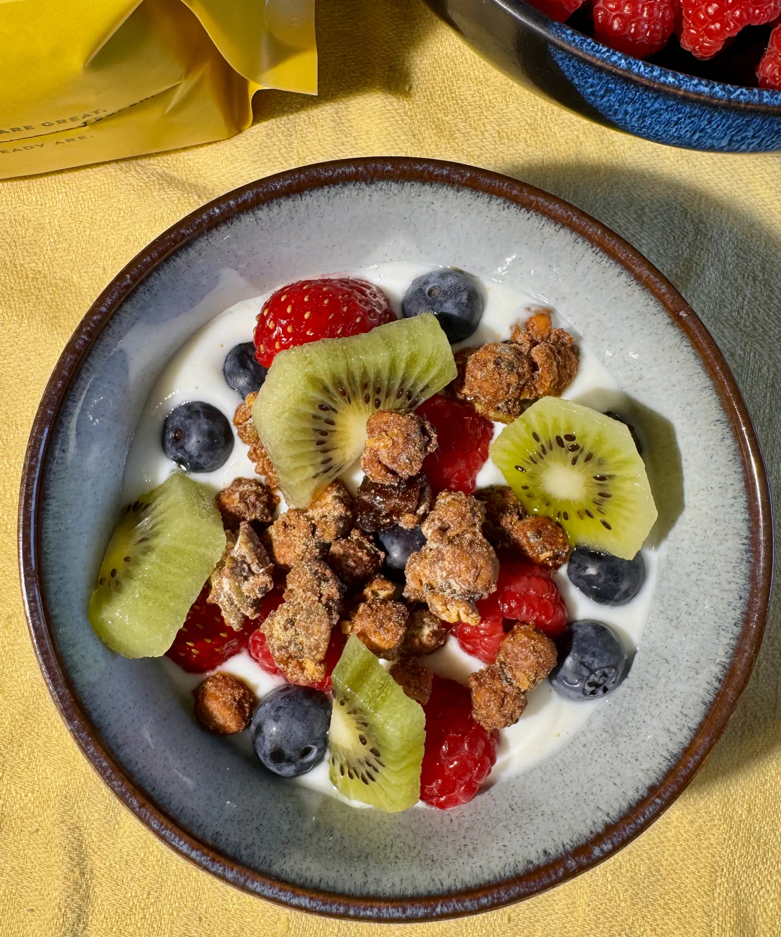 A top-down view of a bowl of Moonbeans granola with yogurt, fresh berries, blueberries, and kiwi slices, on a yellow tablecloth. A yellow bag is partially visible in the upper left, and a blue bowl of raspberries is in the upper right.