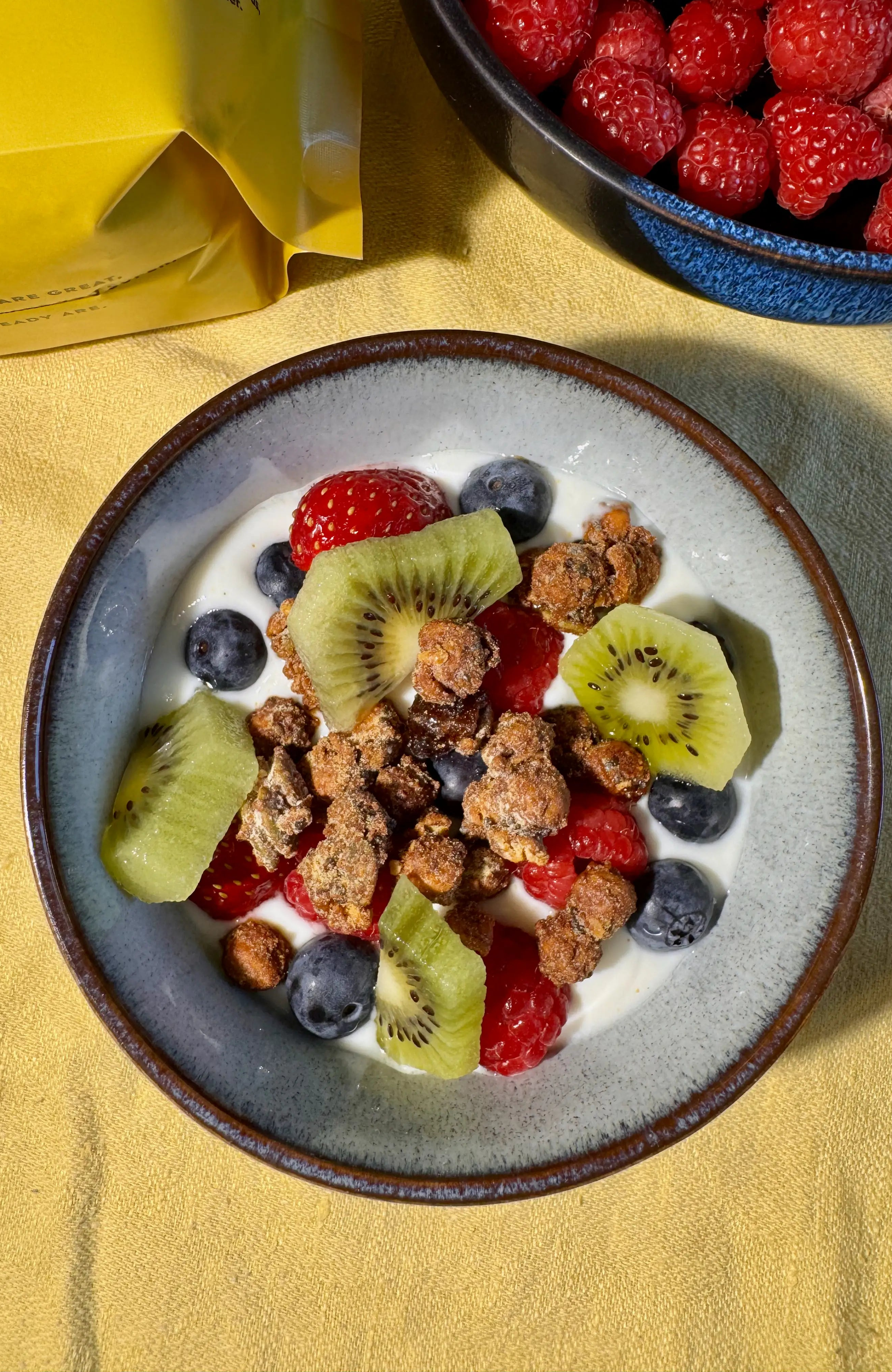 A top-down view of a bowl of Moonbeans granola with yogurt, fresh berries, blueberries, and kiwi slices, on a yellow tablecloth. A yellow bag is partially visible in the upper left, and a blue bowl of raspberries is in the upper right.